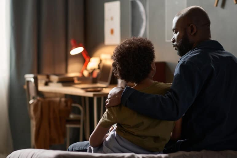 Back view portrait of caring single father embracing son sitting on bed together and talking copy space
